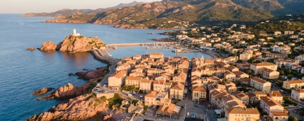 Vue aérienne de l'Île-Rousse en Corse montrant la ville avec ses toits ocre, le phare blanc de la Pietra, les rochers de granite rose et la mer Méditerranée bleue en lumière dorée de fin d'après-midi